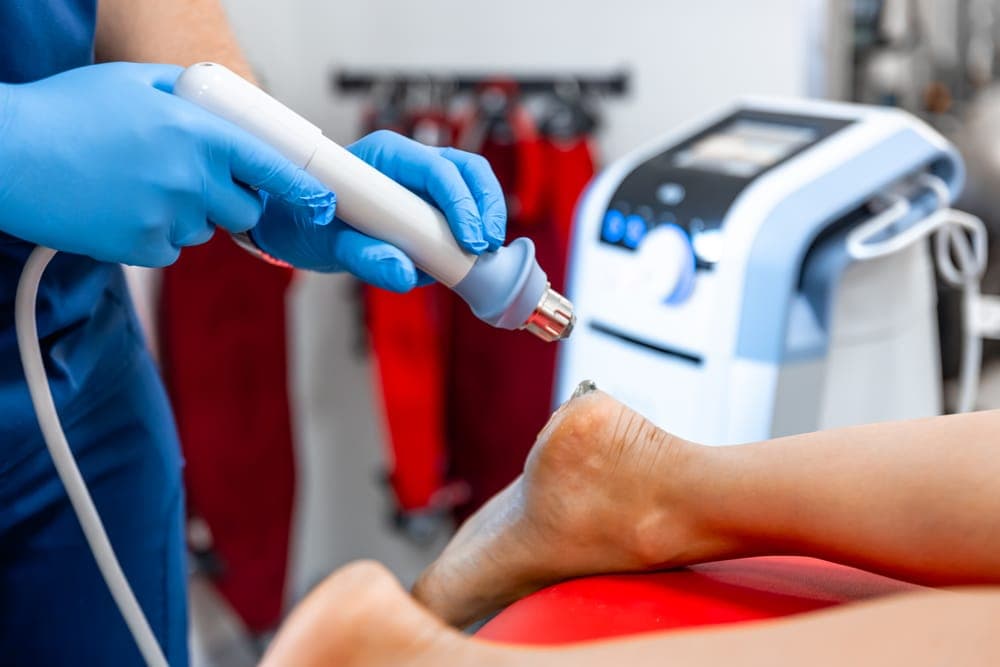 A clinician applying a shockwave therapy handpiece to a patient's heel beside the treatment unit