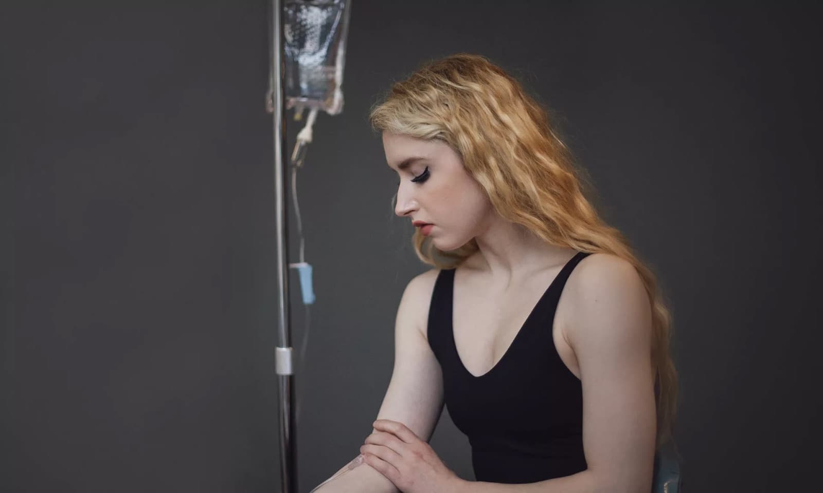 A woman seated beside an IV drip bag hanging from a stand during a vitamin infusion