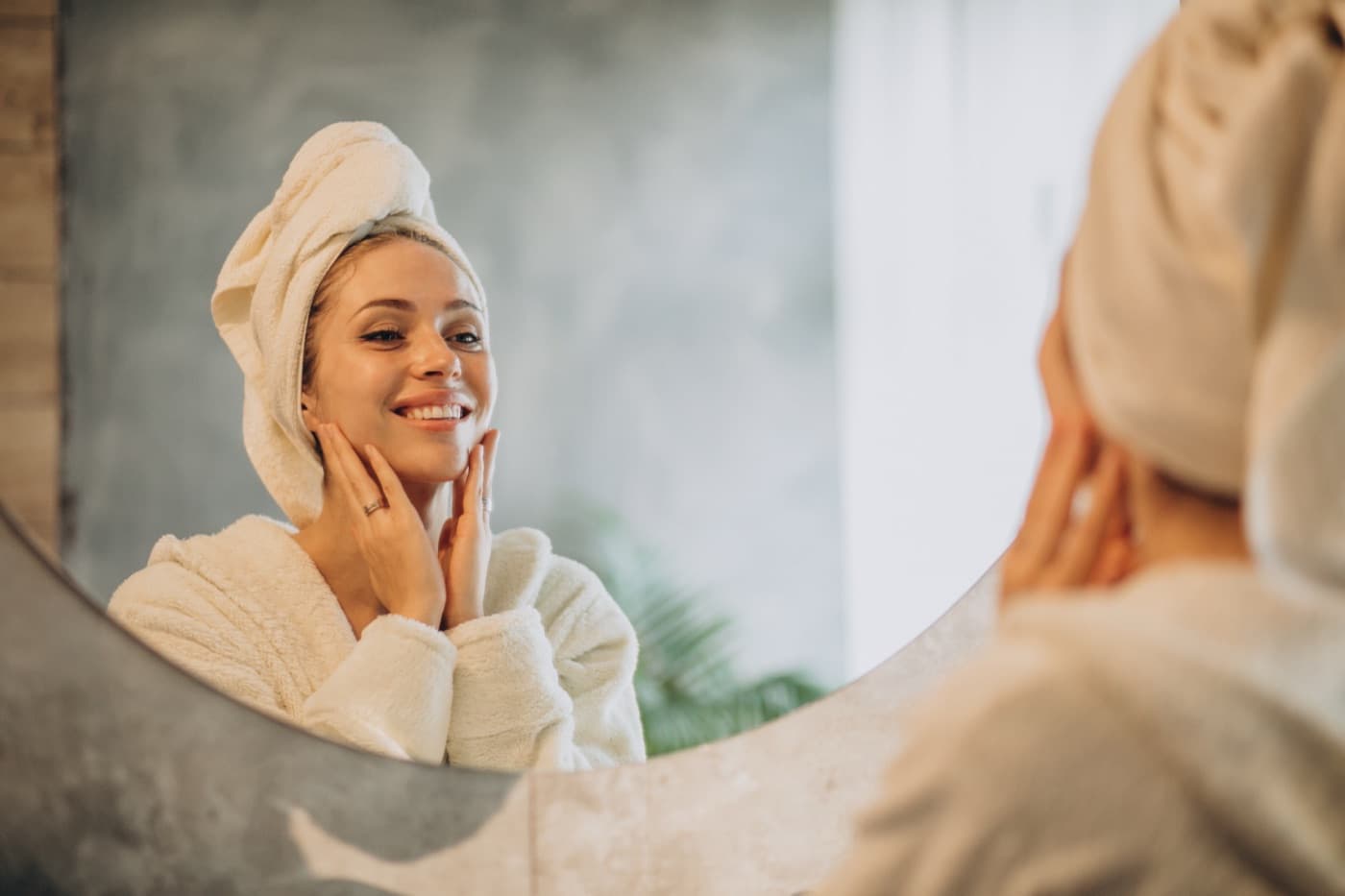 A woman in a robe and hair towel touching her cheek while looking in a mirror