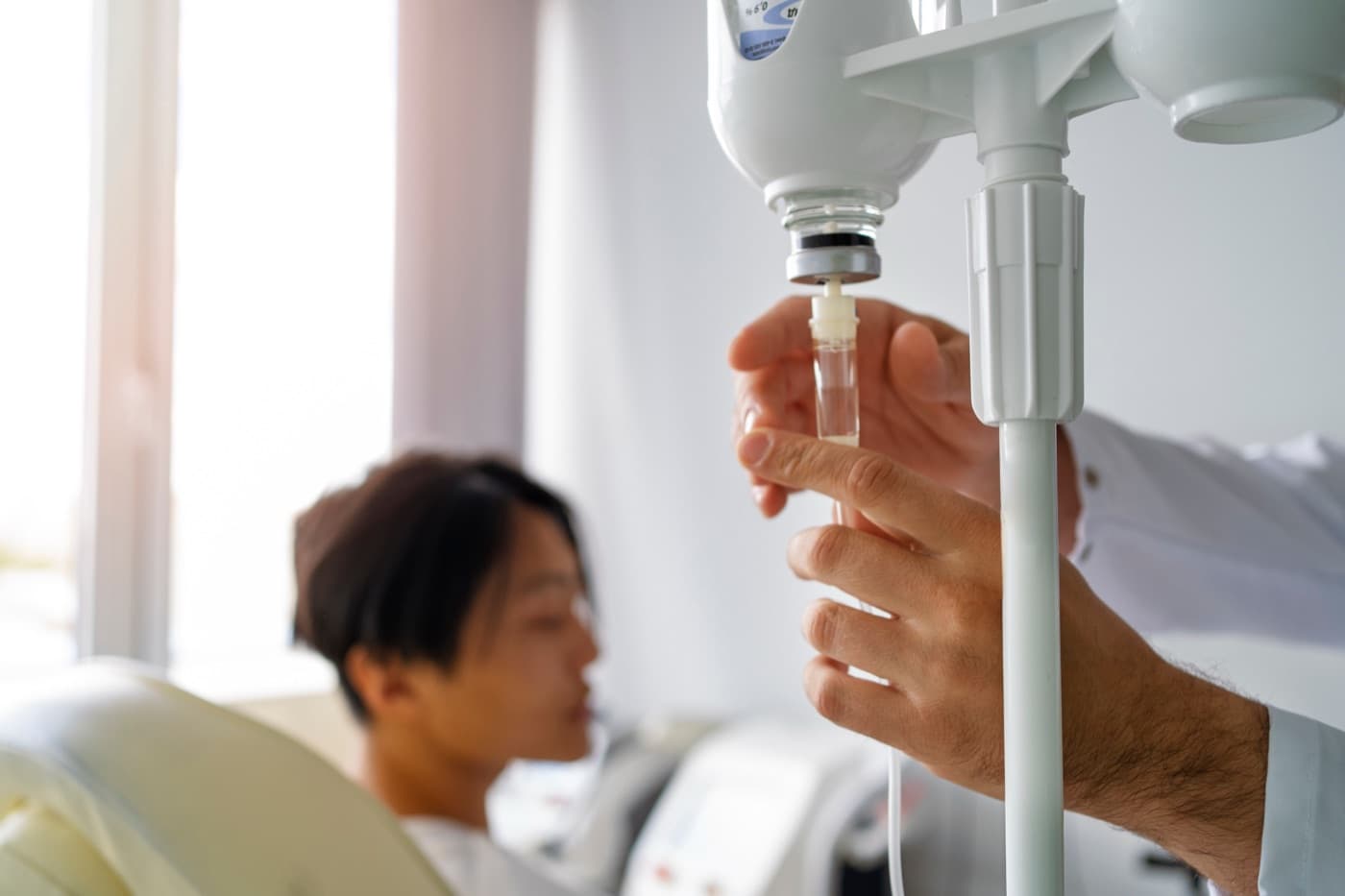 A clinician adjusting an IV drip bag on a stand while a patient rests on a treatment bed by a sunlit window