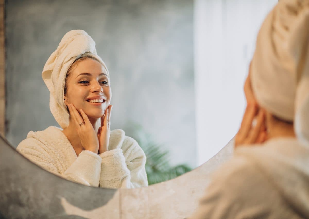 A woman in a robe with a towel wrapped around her hair smiling as she applies cream to her face at a mirror