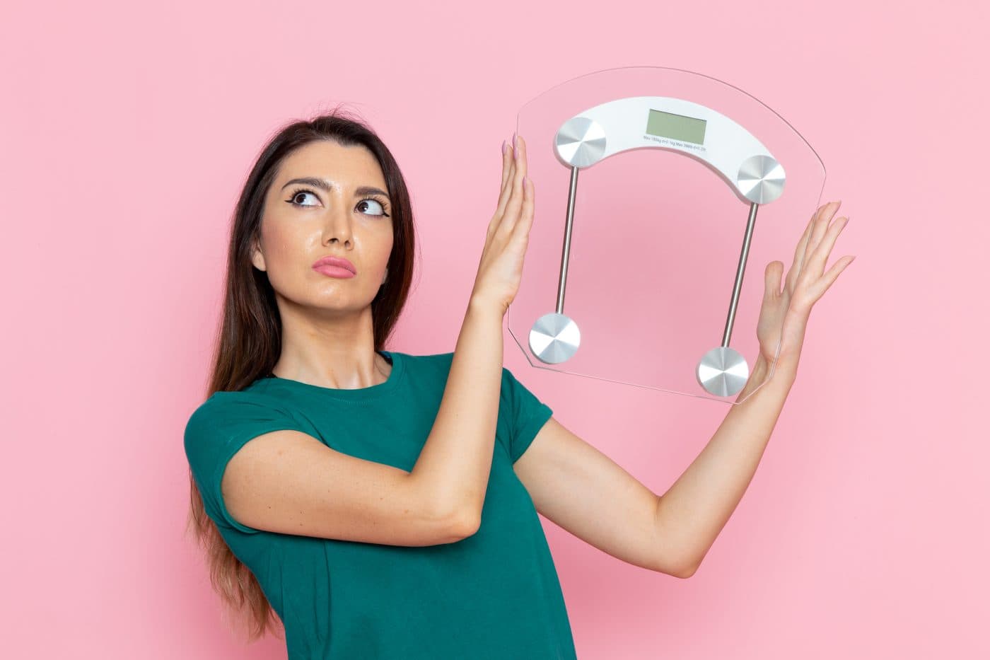 A woman holding a glass bathroom scale at arm’s length with a wary expression, against a pink background