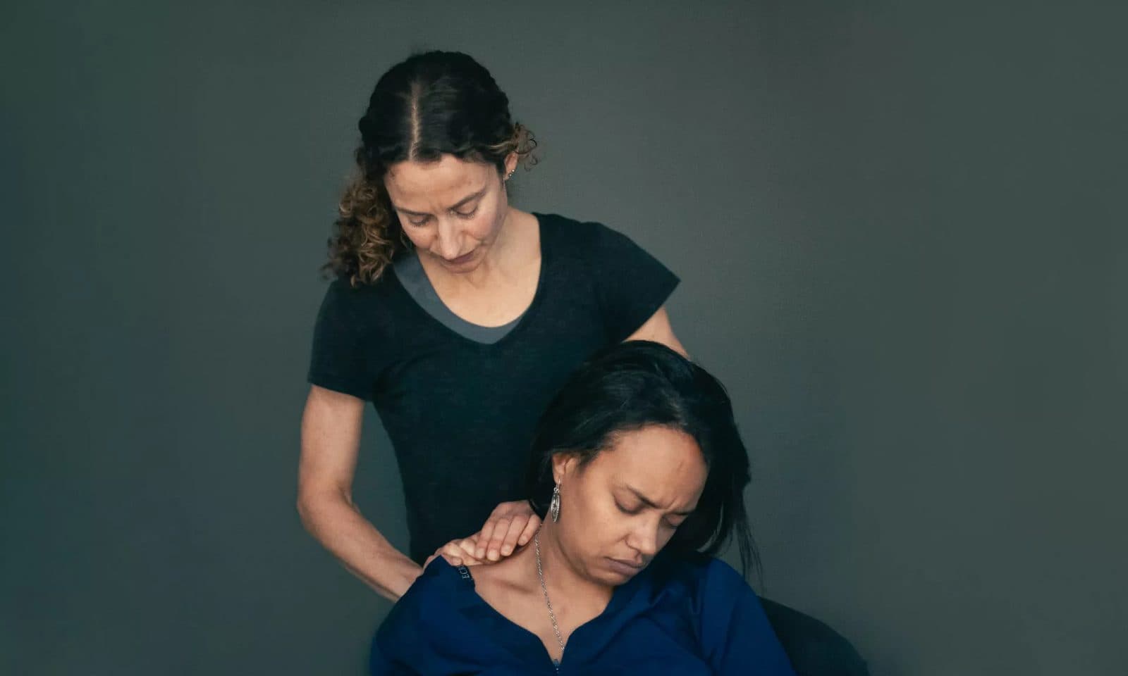 A massage therapist standing behind a seated patient, pressing into the muscles at the base of the patient's neck