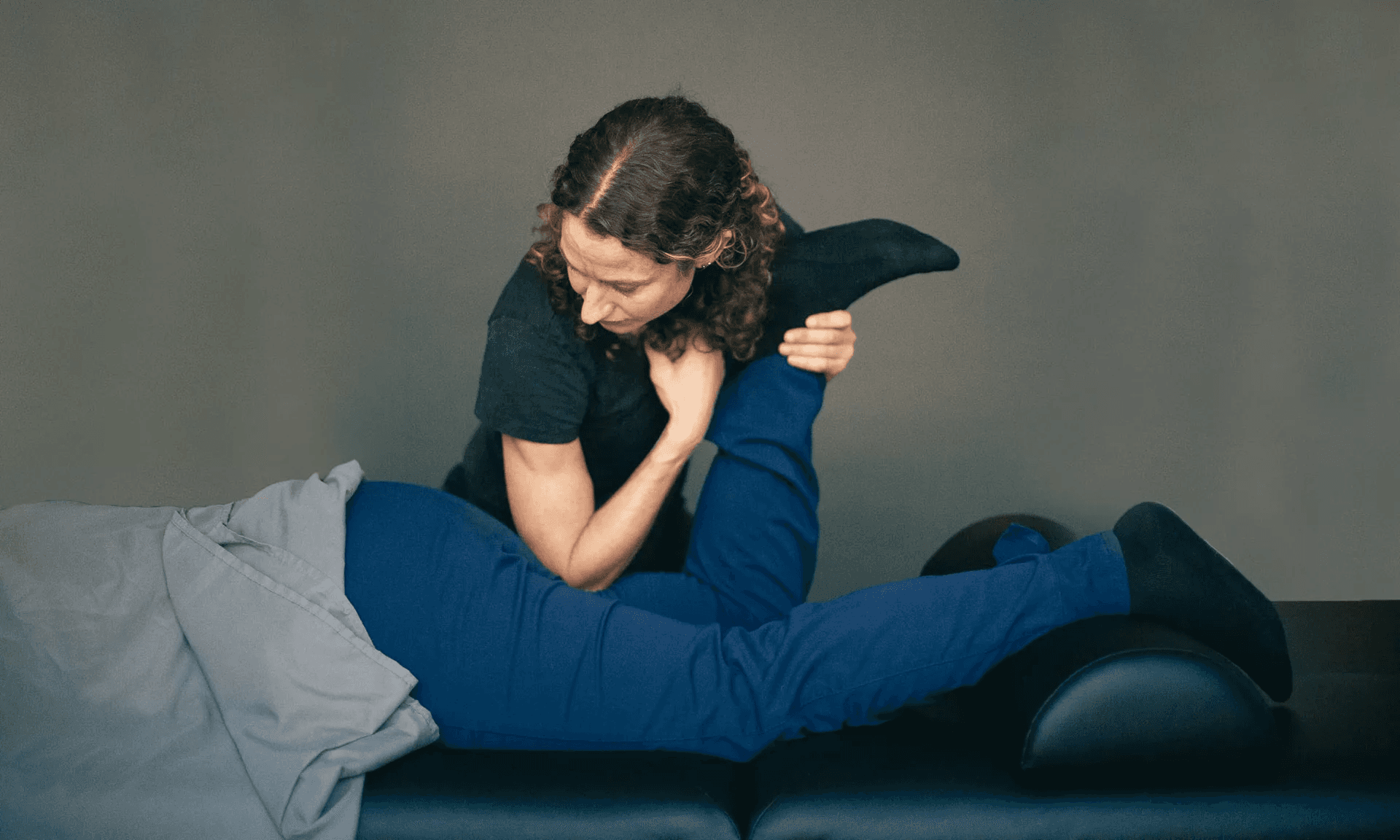 A therapist kneeling beside a side-lying patient on a treatment table, guiding the patient's leg through a supported stretch