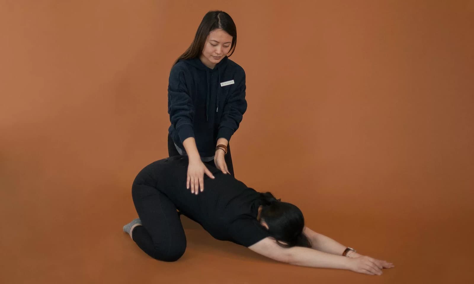 A Physio Logic therapist guiding a patient through a kneeling child's-pose stretch with hands placed on the lower back