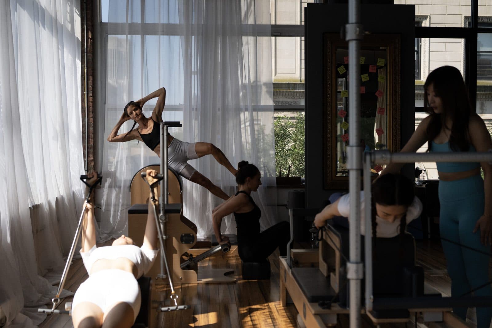 Several clients working on reformers and a ladder barrel in a sunlit Pilates studio while an instructor assists
