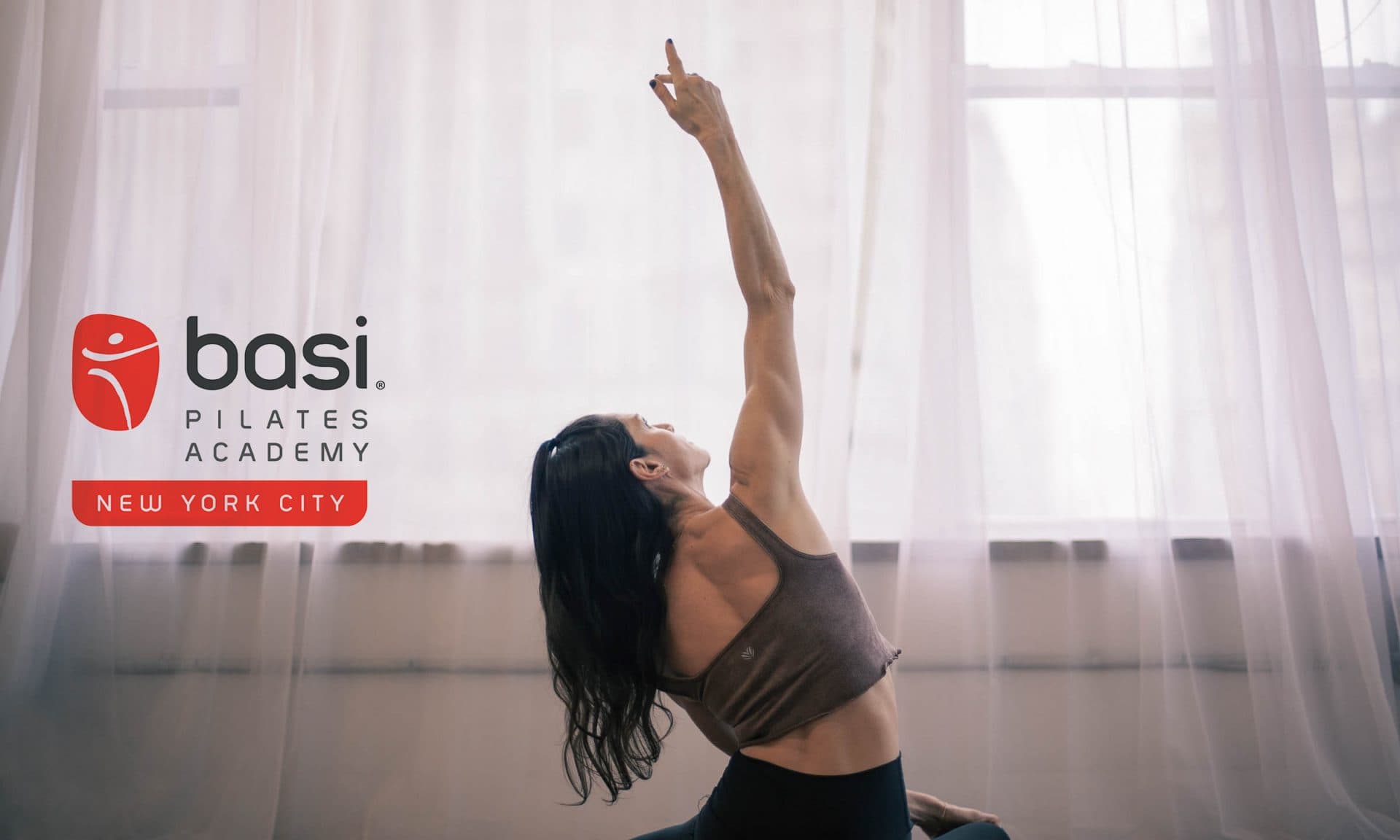 A woman arching back with one arm reaching overhead in a sunlit Pilates studio, beside the BASI Pilates Academy New York City logo